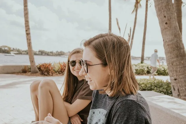 Two young women sitting and talking on a waterfront in the sun.